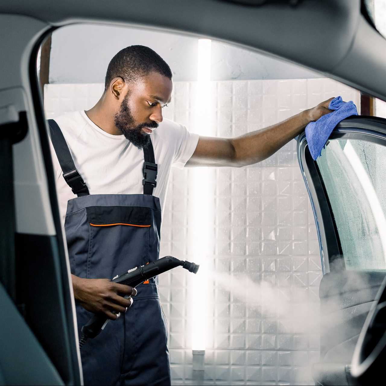 Car detailing, steam cleaning concept. Handsome African man in overalls and t-shirt, worker of car wash center, cleaning car with hot steam cleaner and blue microfiber cloth. Car detailing concept