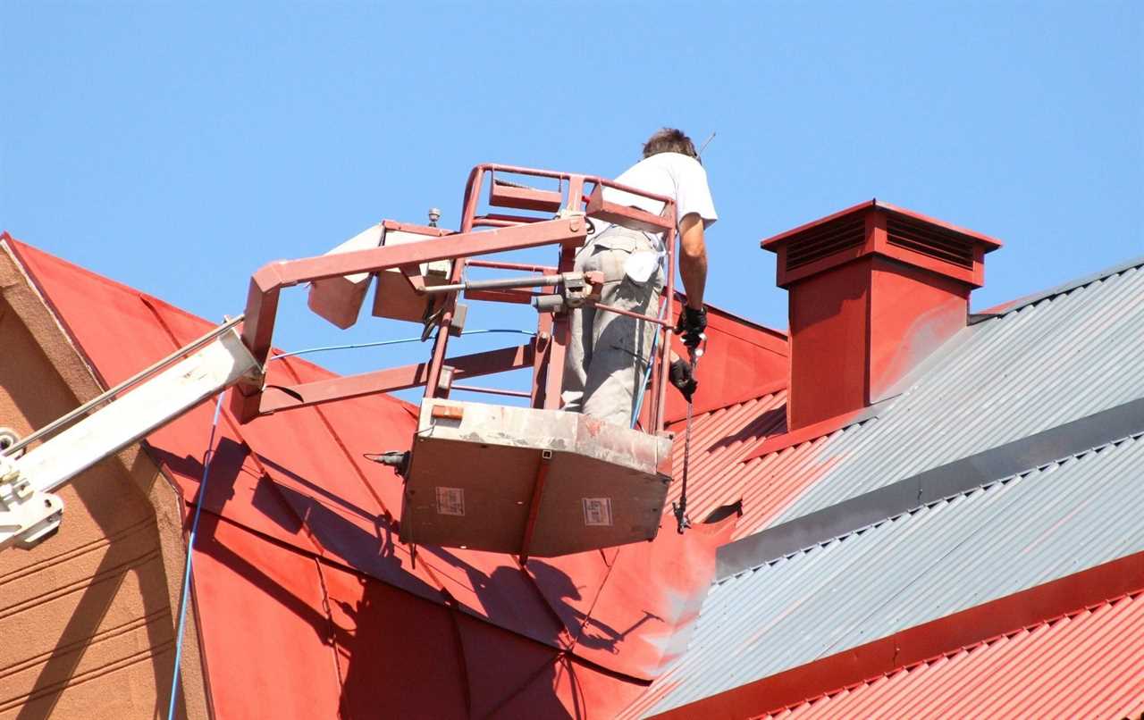 Tradesman Worker Operating a Crane Lift is Painting a Metal Roof on a home Red with a Paint Sprayer in a blue bright day